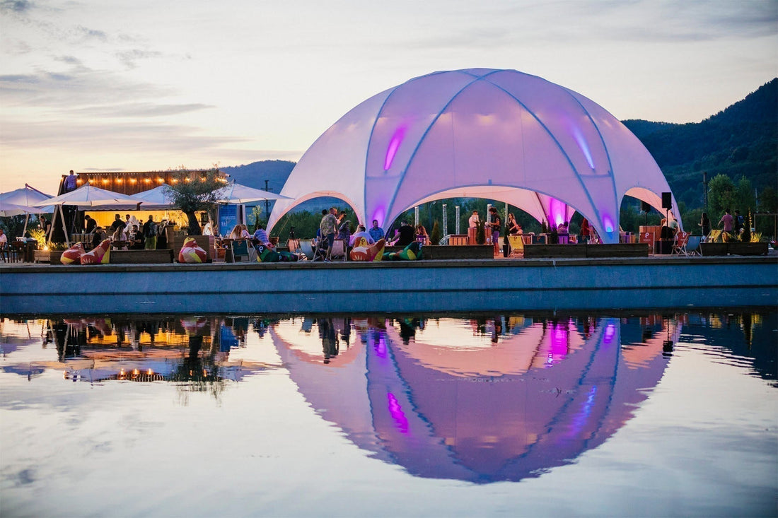 Verlichte Hexadome tent bij zonsondergang, weerspiegeld in het water tijdens een outdoor event.