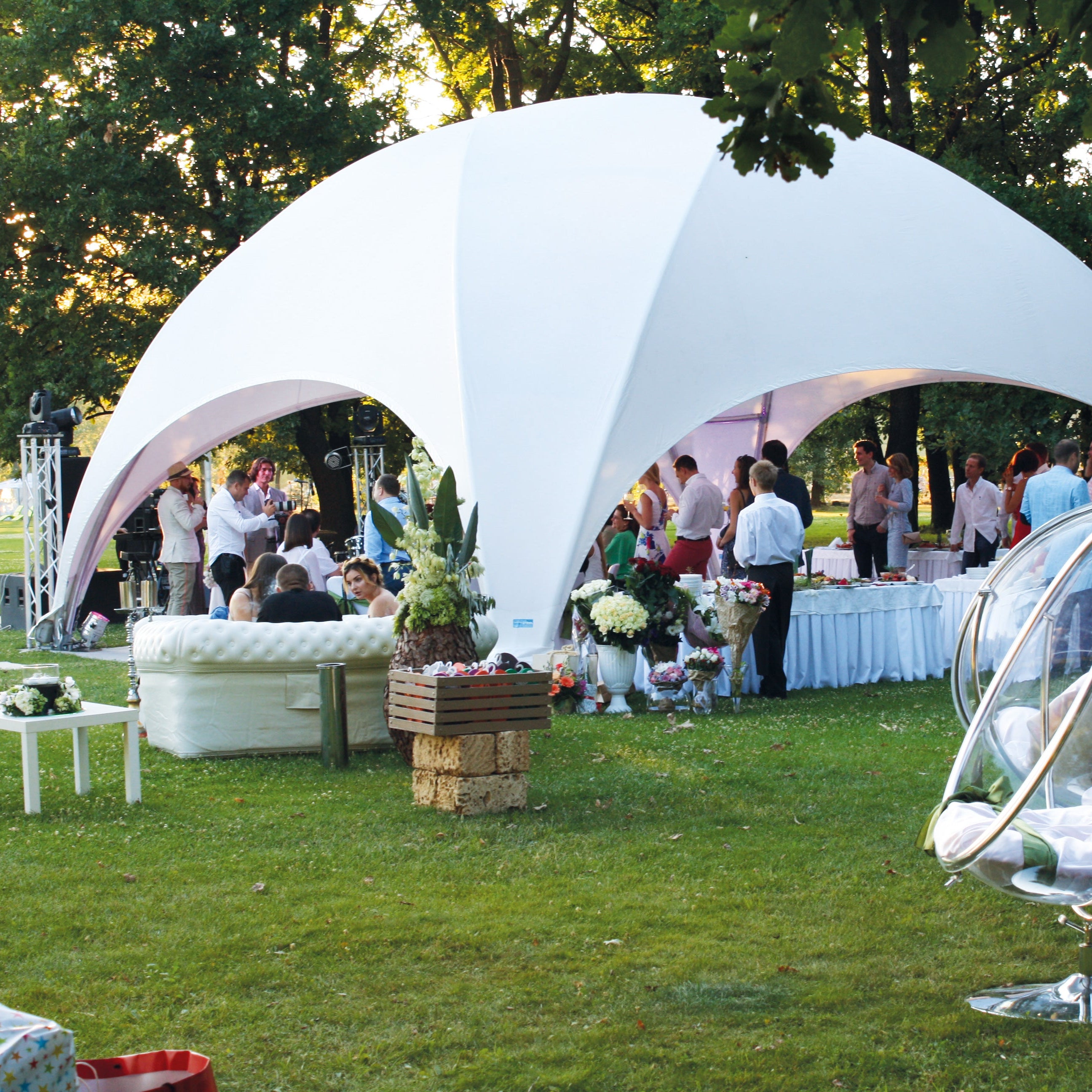 Foto van een bruiloftsopstelling met een witte Crossover tent van Qpparty, geplaatst op een grasveld met gasten, decoratie en lounge-meubilair in een parkachtige omgeving.