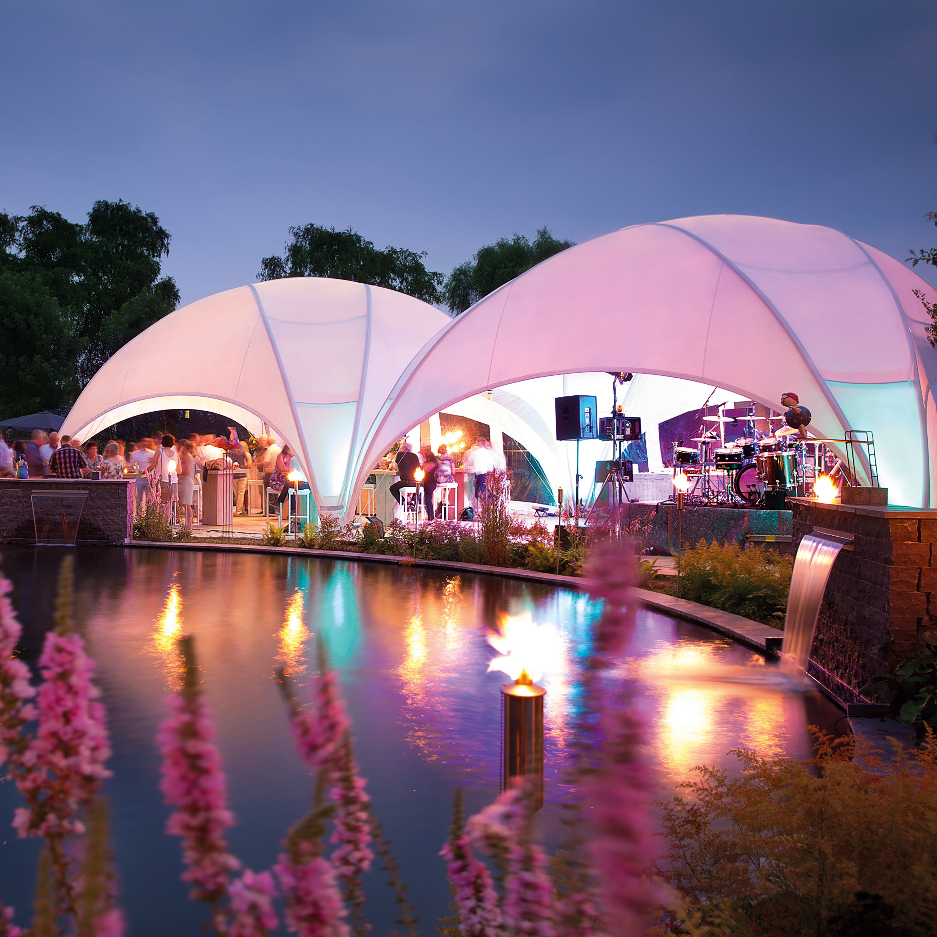 Sfeervolle avondfoto van meerdere verlichte Crossover tenten van Qpparty, opgesteld langs een vijver tijdens een evenement, met kleurrijke verlichting en gasten rondom de tenten.