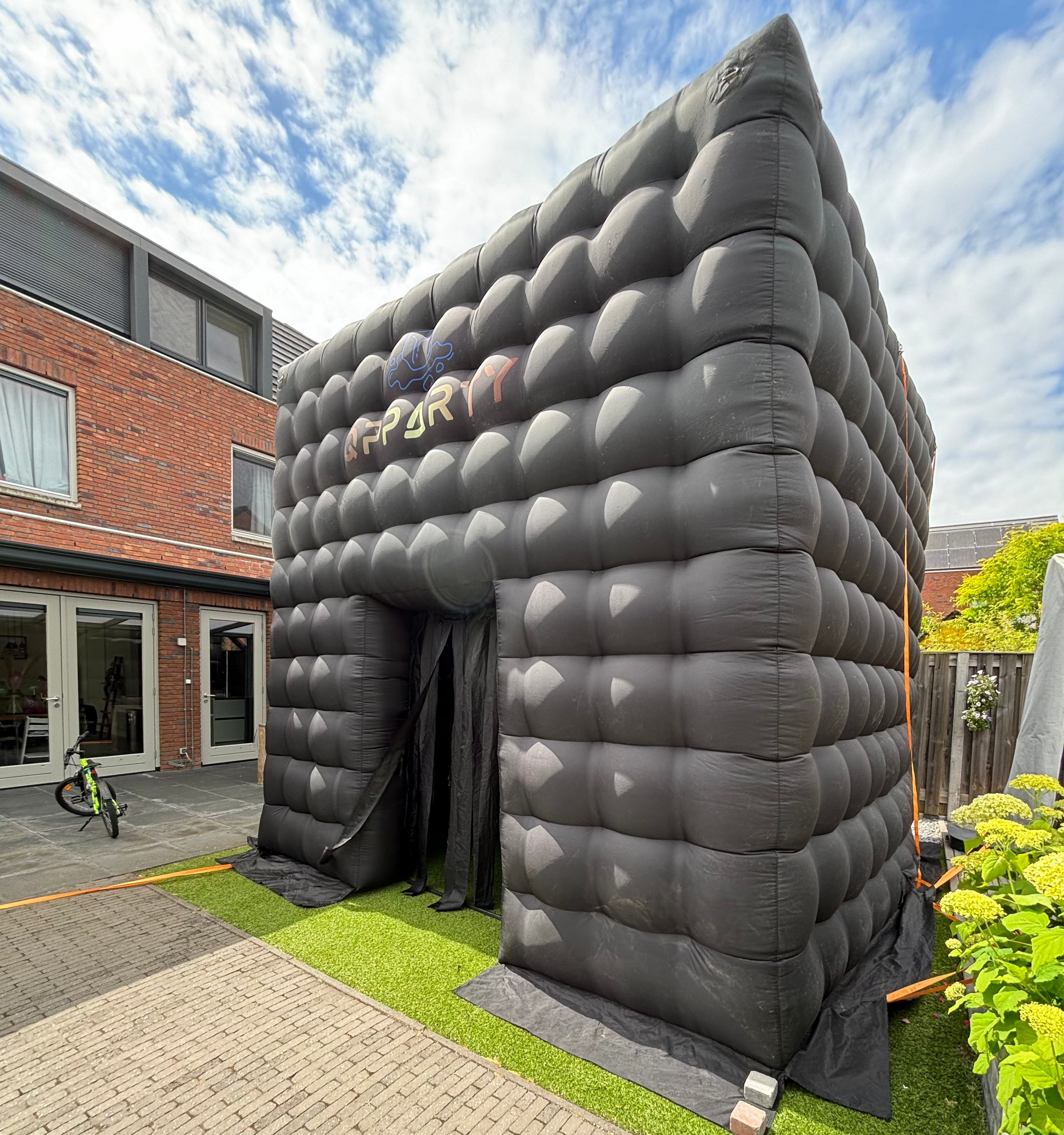 Opblaasbare vierkante Qpparty tent met open ingang, geplaatst op kunstgras naast een woning. Met een capaciteit van 30 tot 40 gasten.