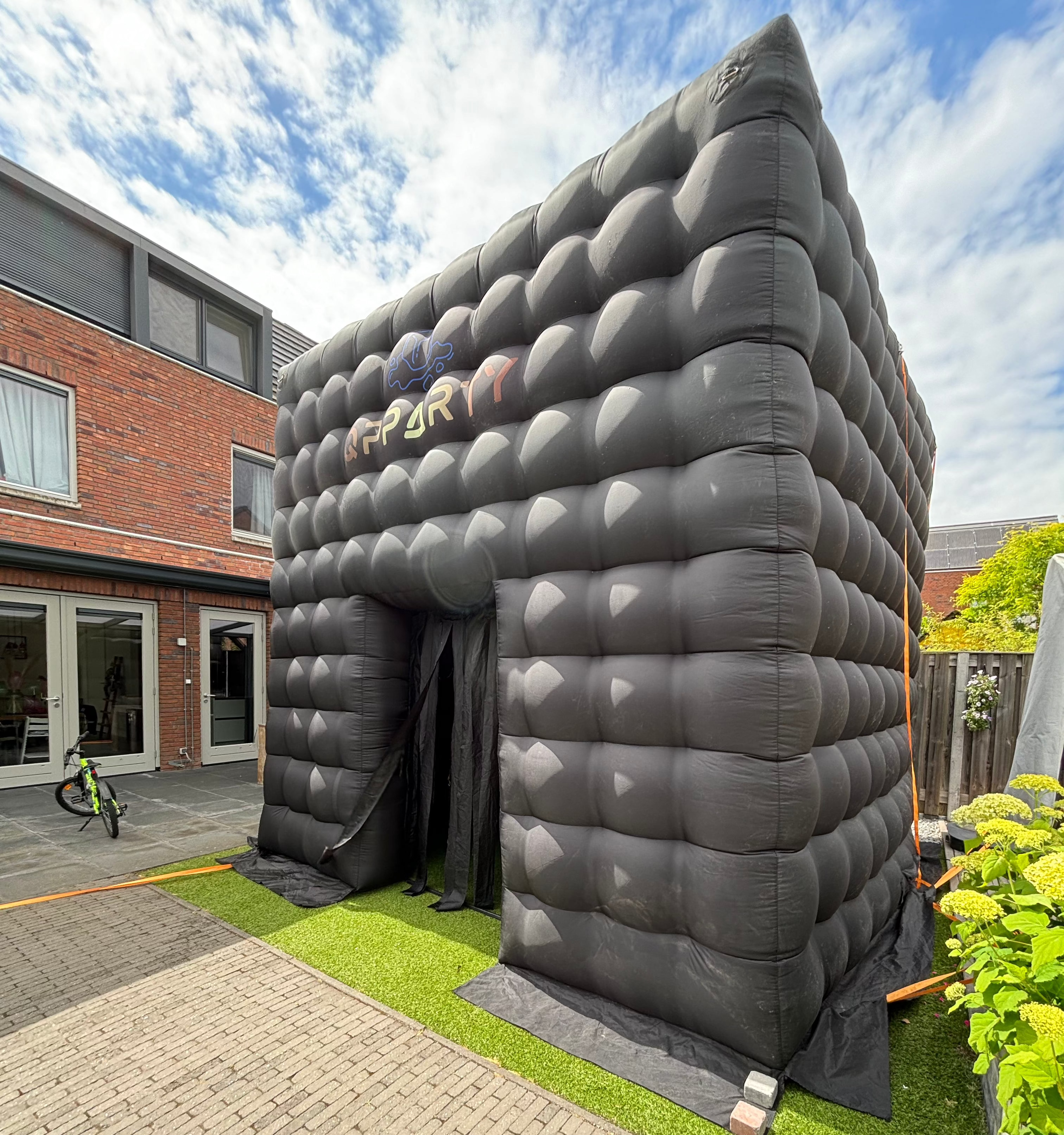 Opblaasbare vierkante Qpparty tent met open ingang, geplaatst op kunstgras naast een woning. Met een capaciteit van 30 tot 40 gasten.