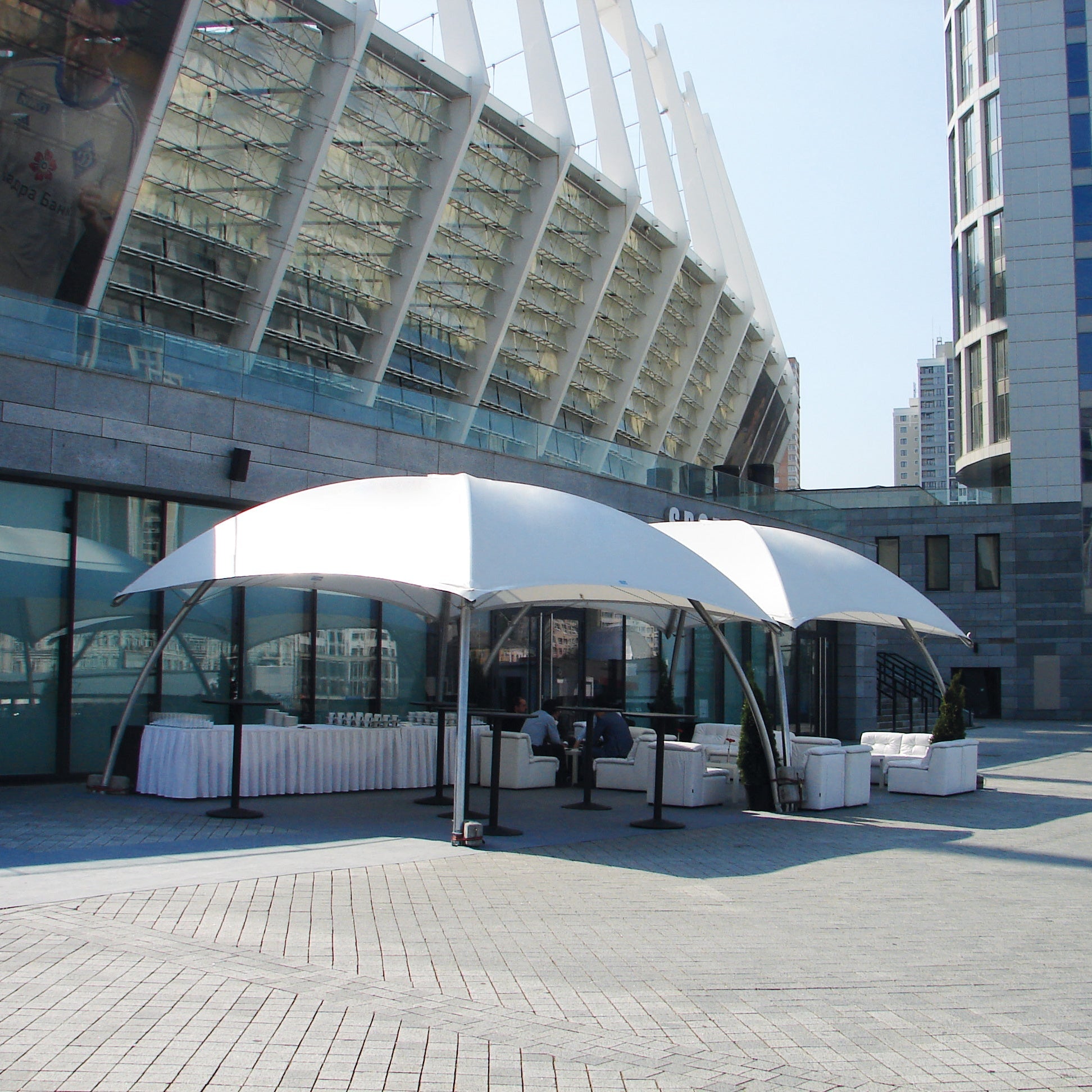 Lounger L met robuust aluminium frame en brandwerende B1-M2 cover, geplaatst op een buitenterras bij een zakelijk evenement, met twee gekoppelde units en staande gasten.