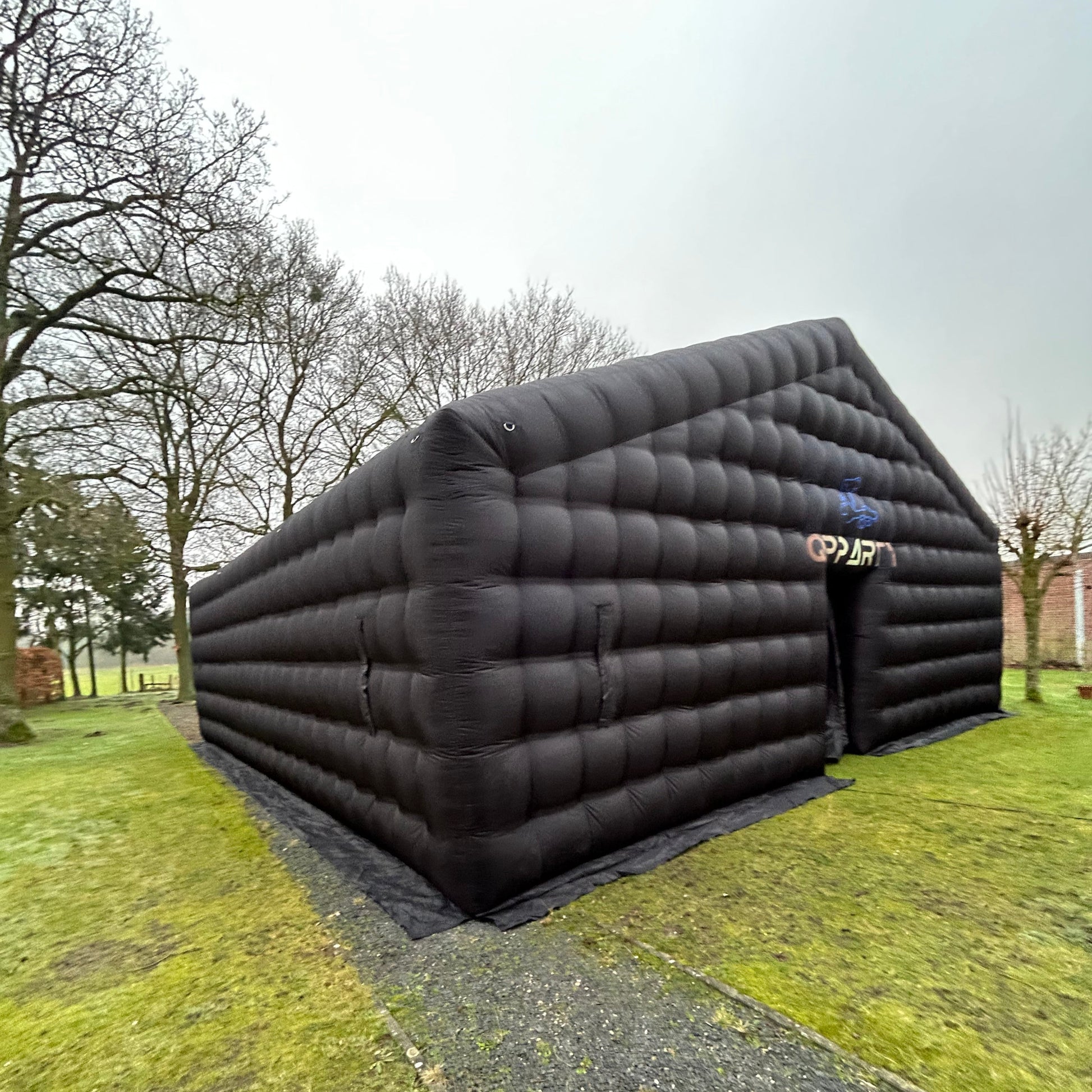 Foto van een mega zwarte Qpparty opblaastent van 4 meter hoog en 10 meter breed en 10 meter lang, opgesteld op een grasveld met zichtbare ingang.