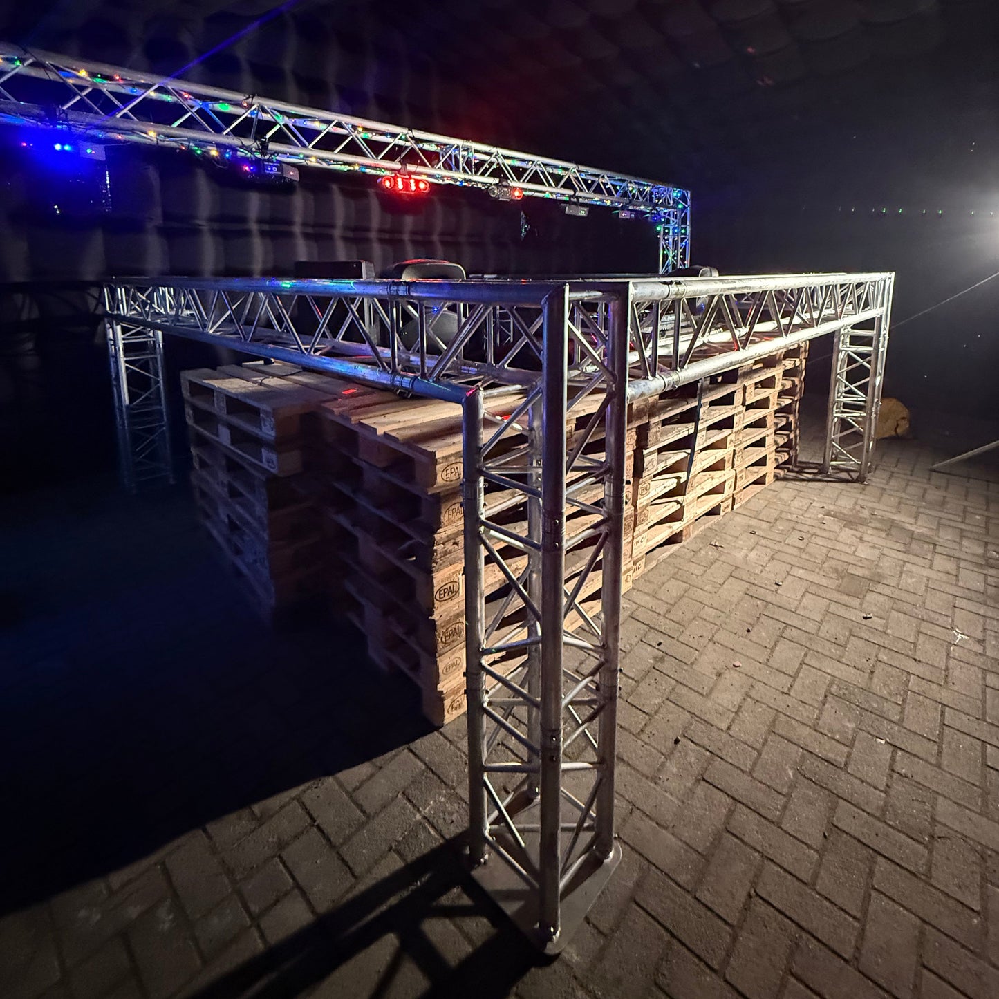 Foto van een QPparty DJ booth opgebouwd uit een trussconstructie van 1,3 meter hoog en 3,6 meter breed en lang, geplaatst boven een palletbar met feestverlichting in een opblaasbare tent.