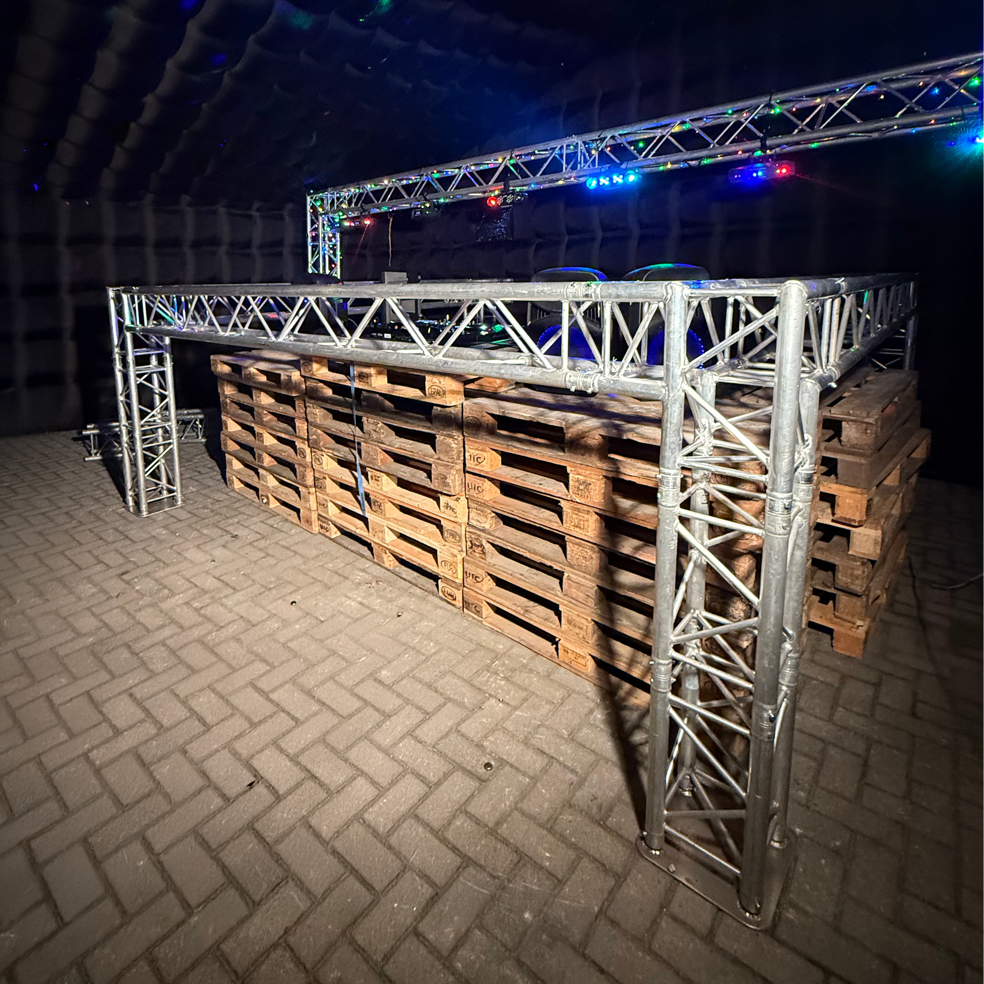 Foto van een QPparty DJ booth opgebouwd uit een trussconstructie van 1,3 meter hoog en 3,6 meter breed en lang, geplaatst in een opblaasbare partytent op een onderstel van houten pallets met feestverlichting op de achtergrond.