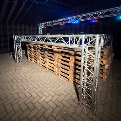 Foto van een QPparty DJ booth opgebouwd uit een trussconstructie van 1,3 meter hoog en 3,6 meter breed en lang, geplaatst in een opblaasbare partytent op een onderstel van houten pallets met feestverlichting op de achtergrond.